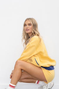 A blonde woman crouching in a studio setting, wearing an oversized yellow sweatshirt with blue side stripes, matching yellow shorts, white socks with red stripes, and white sneakers against a plain white background.